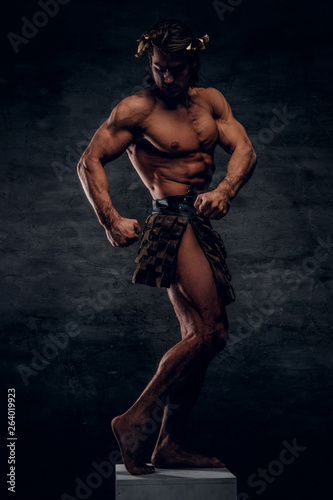 Attractive shirtless man is posing on pedestal and showing his muscules. He is wearing wreath and bandage.