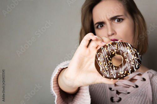 Scared brunette girl with natural makeup holding donut against a grey background. Space for text