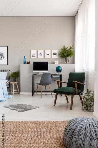 Grey pouf on natural linen carpet in fashionable teenager's room with retro armchair and industrial desk with computer