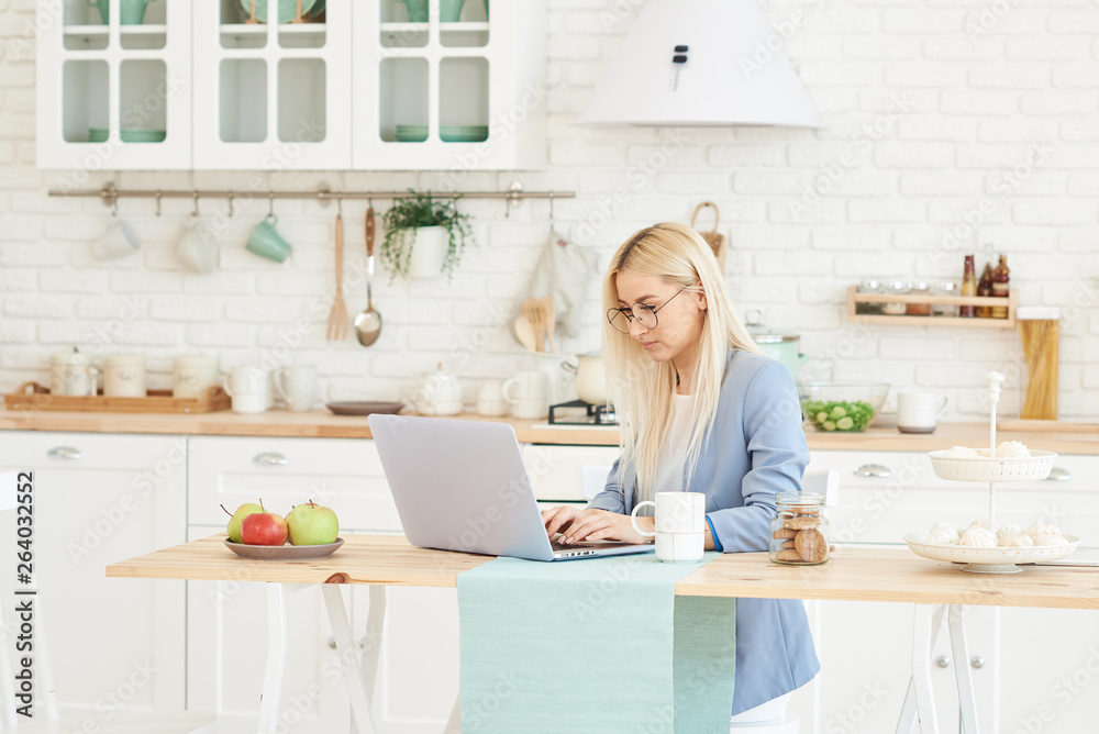 Freelance concept. Beautiful business woman in casual clothes and glasses is examining documents and smiling while working with a laptop in kitchen. Working at home Freelance concept. Beautiful business woman in casual clothes and glasses is examining documents and smiling while working with a laptop in kitchen. Working at home