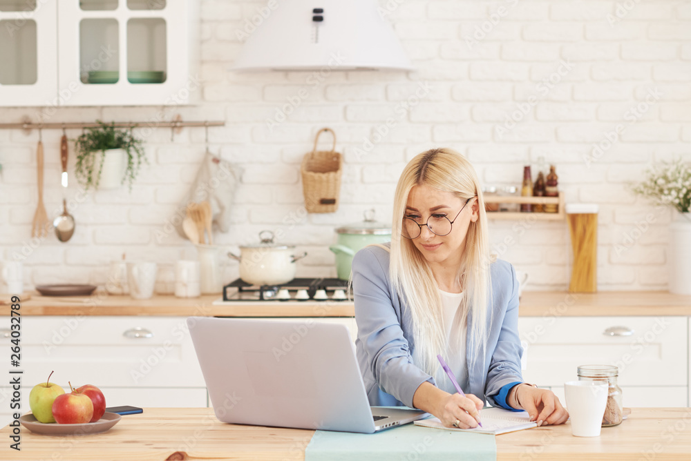Freelance concept. Beautiful business woman in casual clothes and glasses is examining documents and smiling while working with a laptop in kitchen. Working at home Freelance concept. Beautiful business woman in casual clothes and glasses is examining documents and smiling while working with a laptop in kitchen. Working at home