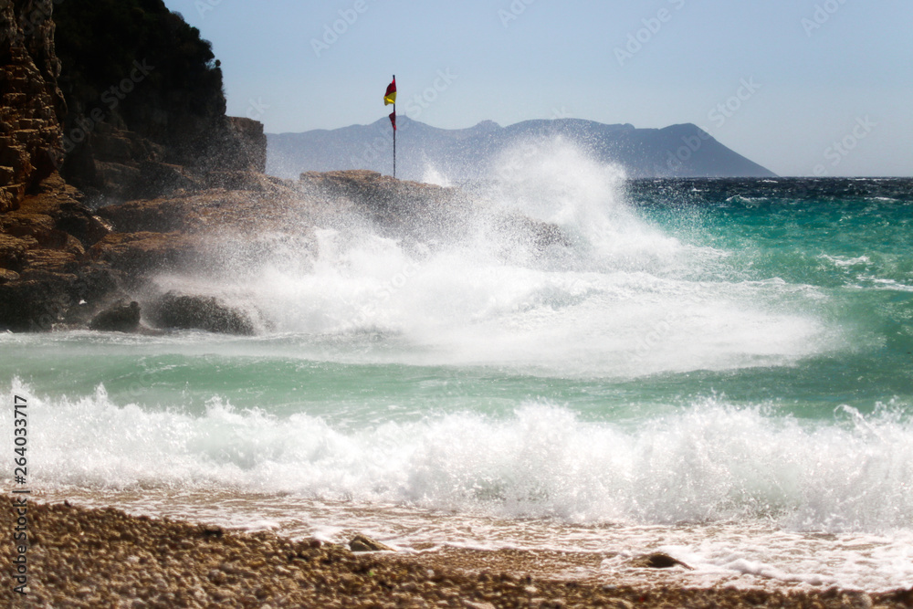 Waves coming from the sea are blasting on the rocks. Water color is turquoise and a red yellow flag is on the rock. Windy and stormy weather at Mediterranean Sea. Buyukcakil Beach, Antalya Turkey