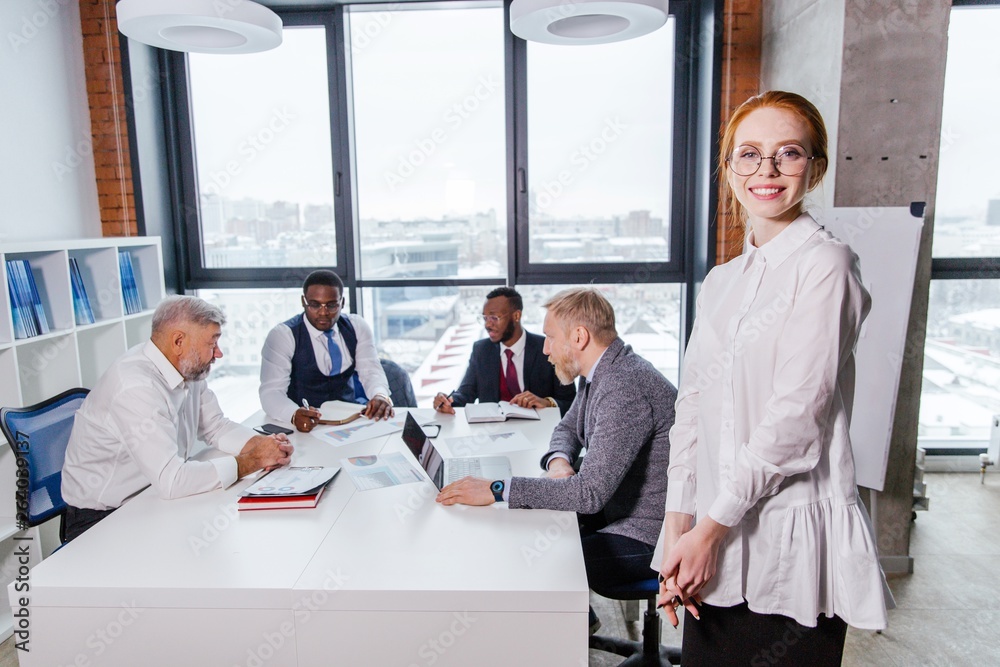 Fototapeta premium Red-haired beautiful girl with glasses standing on the background of business colleagues in a modern office