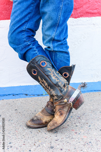 Old cowboy boots are weathered and brown with stitched design on the side with spurs worn by an old cowboy in jeans.