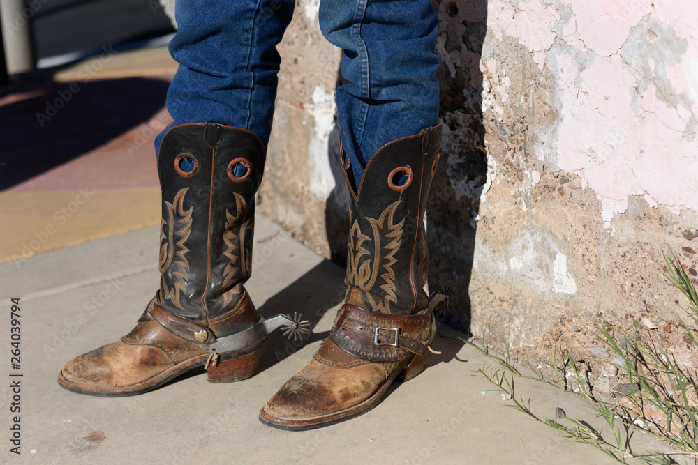 Old cowboy boots are weathered and brown with stitched design on the side  with spurs worn by an old cowboy in jeans. Stock Photo | Adobe Stock