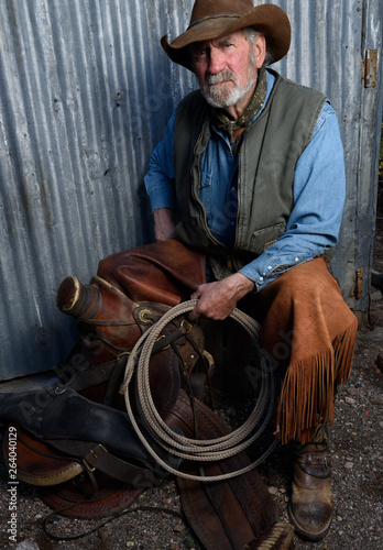 Old cowboy with gray beard wears leather hat, leather chaps, and holds a lasso and is sitting with a leather saddle.