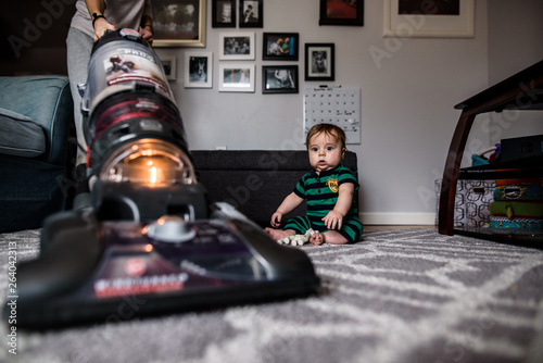 startled baby watches as mother vacuums the rug he is sitting on