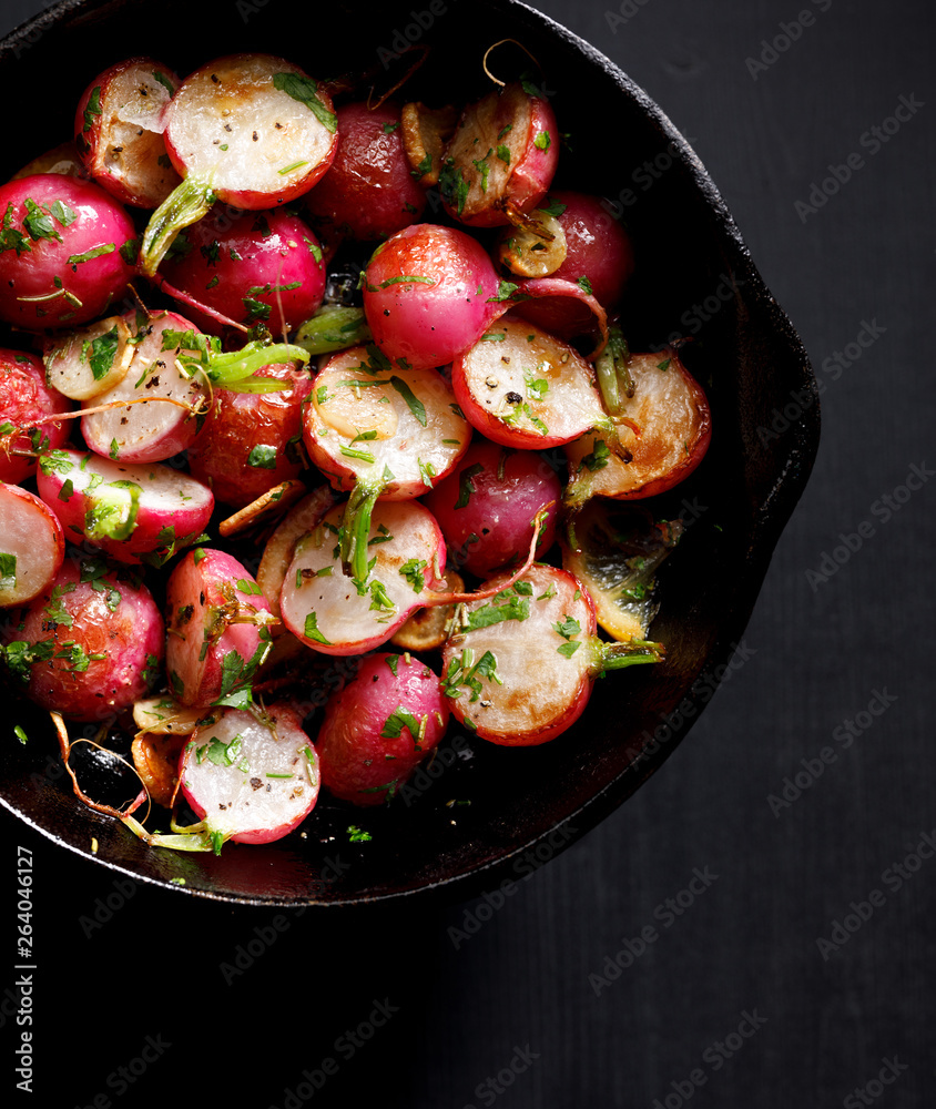 Fried radishes with the addition of herbs and spices on a cast-iron pan on a black background, top view., close-up Vegan food