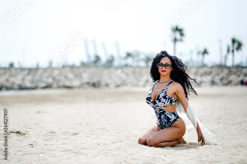 Outdoor fashion photo of beautiful sensual girl with dark curly hair wearing swimsuit, posing on the beach