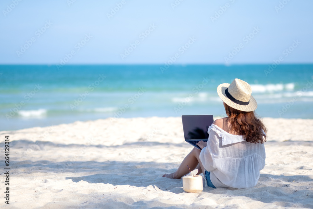 A woman using and typing on laptop computer keyboard while sitting on a beautiful beach