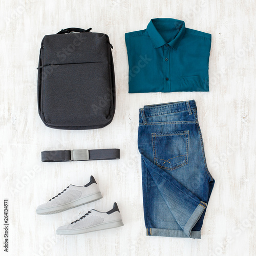 Denim shorts, blue shirt, light gray sneakers, dark grey backpack and belt. Overhead view of men's casual summer outfits on white wooden background. Flat lay, top view. Man outfit.