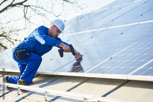 Man worker in blue suit and protective helmet installing stand-alone solar panel system. Professional electrician mounting solar module on roof of modern house. Alternative energy ecological concept.