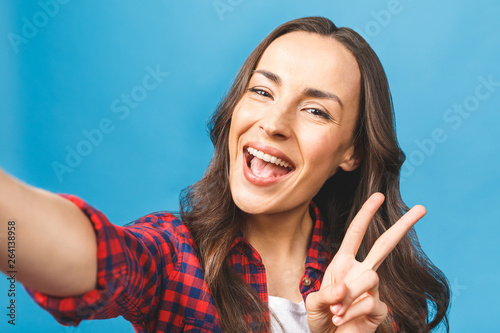 Happy woman in glasses with beaming smile making selfie and showing v-sign. Isolated against blue background.