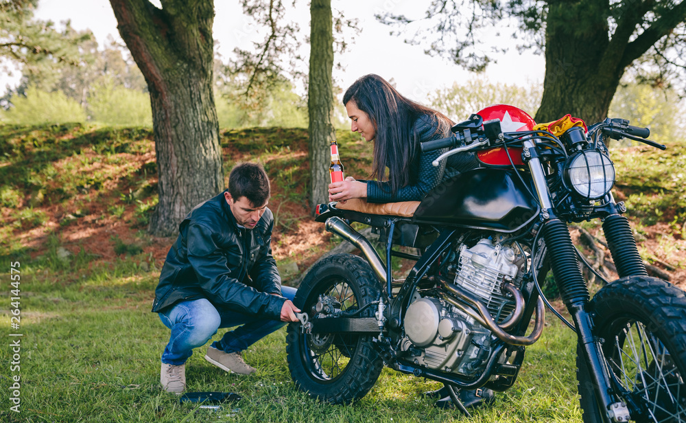 Young man fixing his damaged motorcycle while his girlfriend looks at him taking a beer outdoors