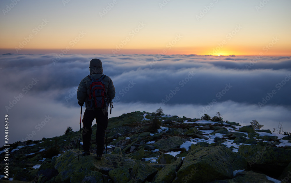 Back view of tourist in warm jacket with backpack and trekking sticks on rocky mountain peak on background of valley covered with white clouds stretching to horizon and bright morning sky at sunrise.