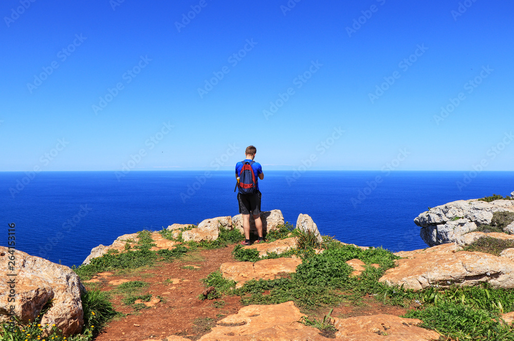 Adult traveler stands on the edge of cliff and takes some picture. Younger, blue sky, blue sea. Human is on the top of hill and enjoying view to down on mediterranean sea in Cyprus nearby Poli city