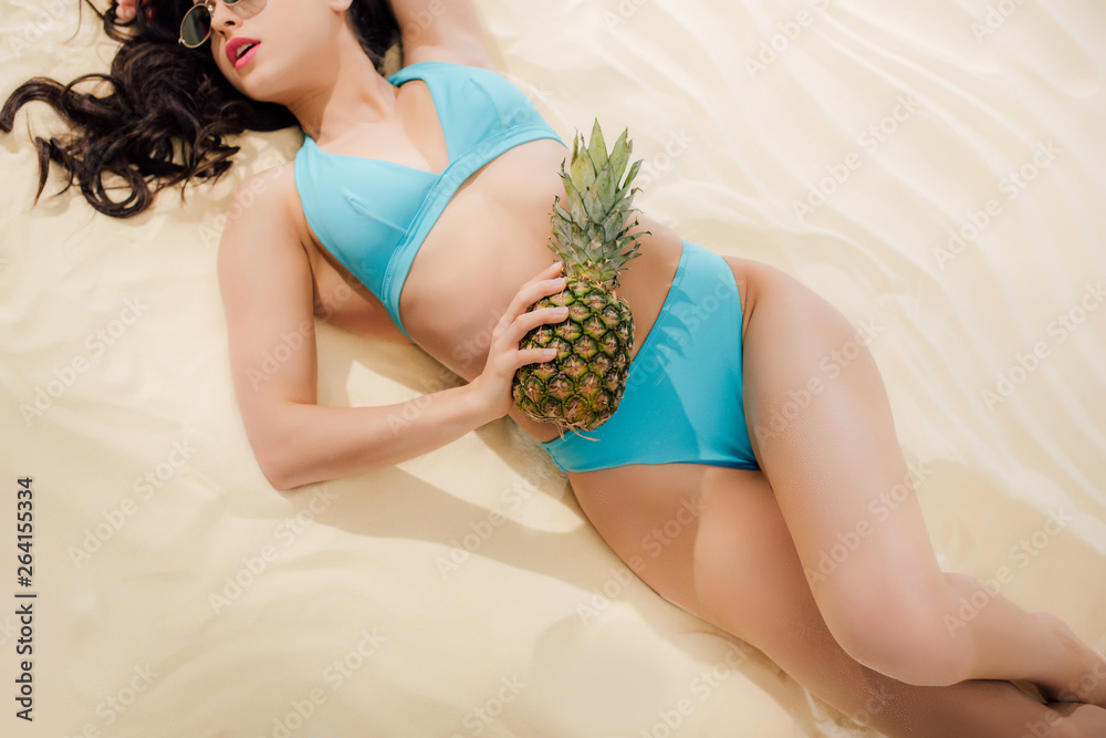 partial view of sexy girl in blue Bikini posing with pineapple while lying  on sandy beach Photos | Adobe Stock