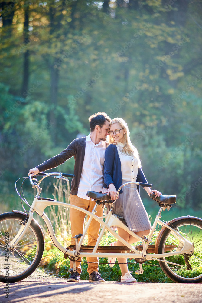 Obraz premium Young happy couple tourists, bearded man and attractive woman close together at tandem double bicycle outdoors in summer park or forest on blurred sunny green trees dense foliage background.