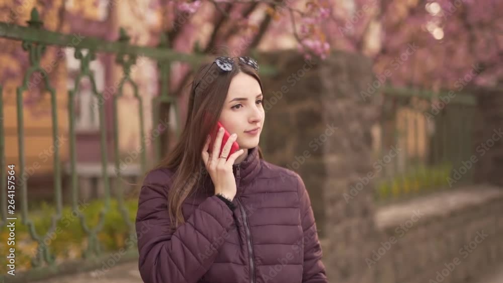 Young Caucasian Female Standing on Street and Nervously Talking with Somebody With Smartphone. Blurred Background Young Caucasian Female Standing on Street and Nervously Talking with Somebody With Smartphone. Blurred Background