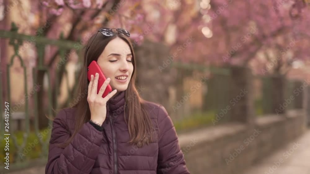 Young Happy Caucasian Female Standing on Street and Talking With Somebody on Smartphone and Smiles. Blurred Background and Light Camera Movement Young Happy Caucasian Female Standing on Street and Talking With Somebody on Smartphone and Smiles. Blurred Background and Light Camera Movement