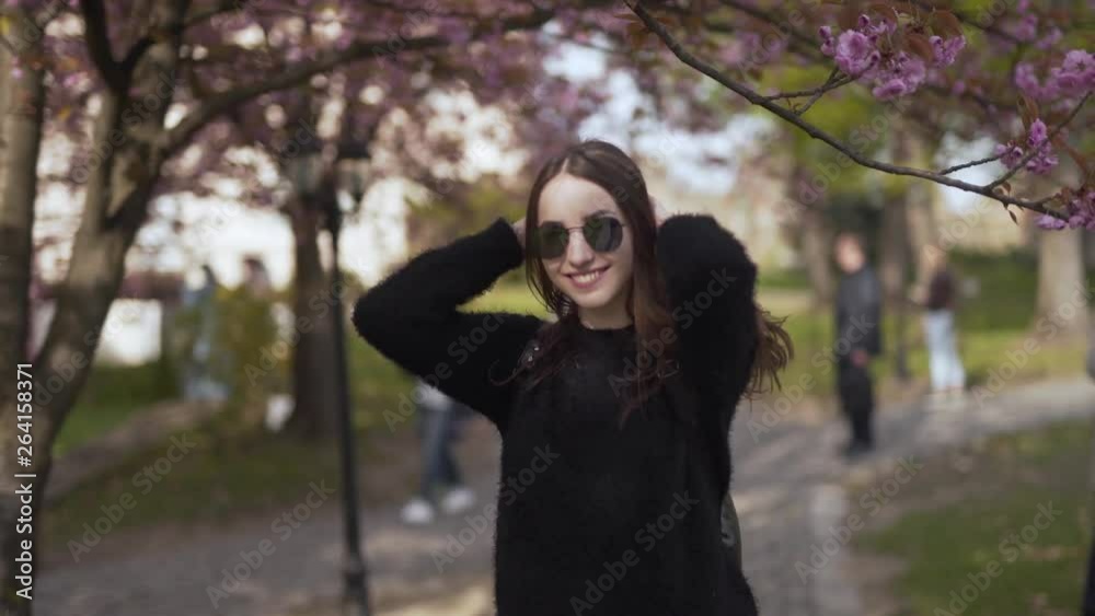 Young Caucasian Female With Sunglasses and Backpack Spinning Around in Front of Camera Under Sakura Cherry Blossom Tree on Spring Day. Blurred Background Young Caucasian Female With Sunglasses and Backpack Spinning Around in Front of Camera Under Sakura Cherry Blossom Tree on Spring Day. Blurred Background