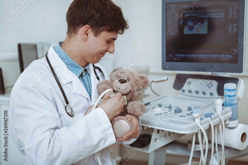 Handsome cheerful male doctor using ultrasound scanner on a plush toy teddy bear, copy space. Charming friendly pediatrician using ultrasound machine at his office. Children, health concept photo