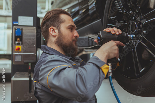 Handsome bearded car mechanic servicing an automobile at his workshop, torquing the lug nuts. Attractive male technician in grey uniform repairing wheels of a SUV vehicle. Safety, insurance concept