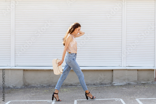 Young stylish woman wearing beige cami silk top, blue cropped denim jeans,  black high heel sandals and holding clutch bag walking in the city street.  Trendy casual outfit. Street fashion. Stock Photo