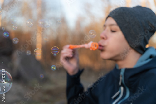 teen boy in blue jacket and grey hat blowing bubbles in Park