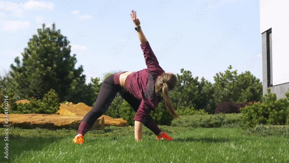 Slender and beautiful young woman in sportswear preparing for workouts on the lawn near her house, doing exercises for warm-ups. Young girl athlete stretches on a meadow with green grass in the park Slender and beautiful young woman in sportswear preparing for workouts on the lawn near her house, doing exercises for warm-ups. Young girl athlete stretches on a meadow with green grass in the park