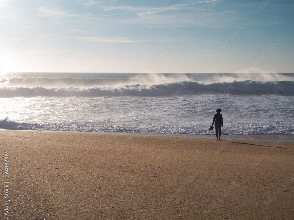 A young woman walking along the wide beach with dangerous curving waves; stormy sea in clear weather Nazare; leisure activity; boots in hand; crystal clear drops of seawater fly in the air; Portugal