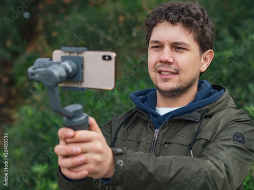 A young man making photo and video of beautiful locations with stabilizer for mobile phone; Smartphone attached to a gimbal; touristic park on background; new technology for travelers; compact devices