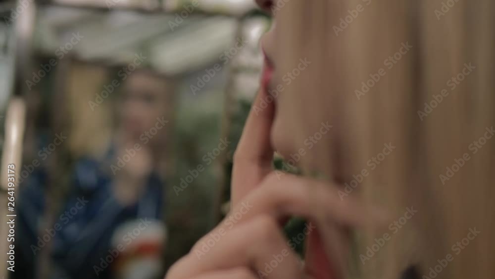 Beautiful blue-eyed blonde admires herself in mirror. She is in botanical garden greenhouse wear striped blue jacket, touching your fingers to your lips. In the background is pink wall and green trees Beautiful blue-eyed blonde admires herself in mirror. She is in botanical garden greenhouse wear striped blue jacket, touching your fingers to your lips. In the background is pink wall and green trees