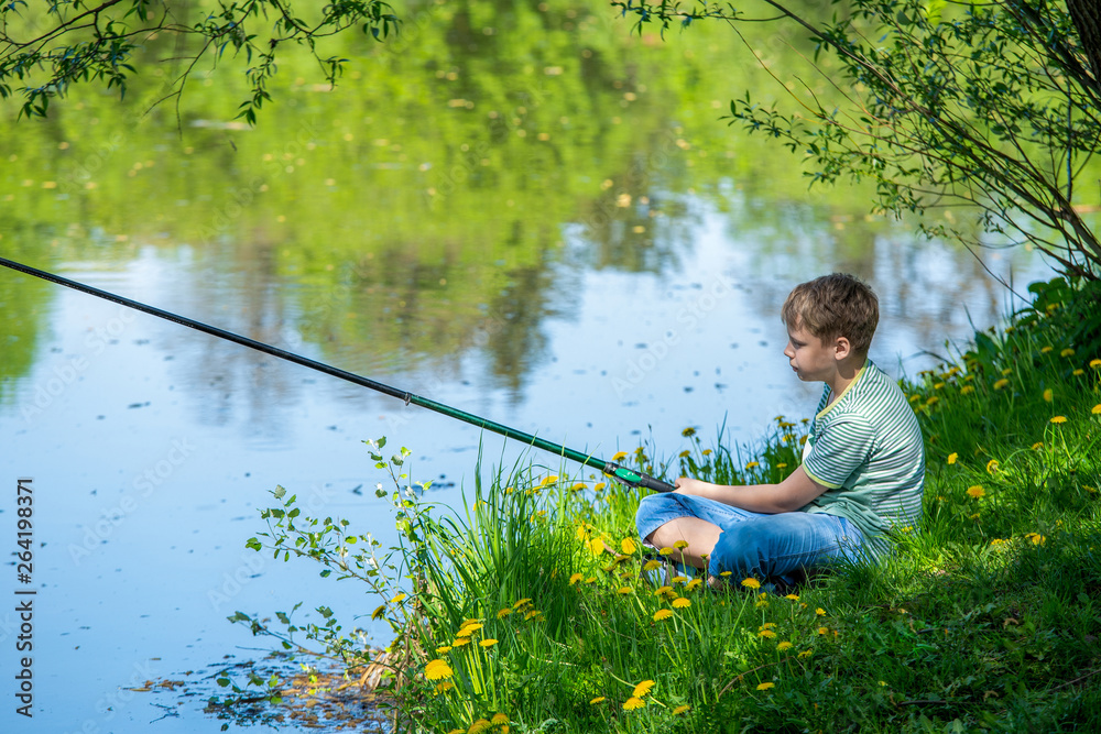 Young kid fishing alone on river in scenic place during summer vacation in  countryside. Horizontal color photography of child sitting in scenic  beautiful landscape at grassy green hill under old tree. Stock