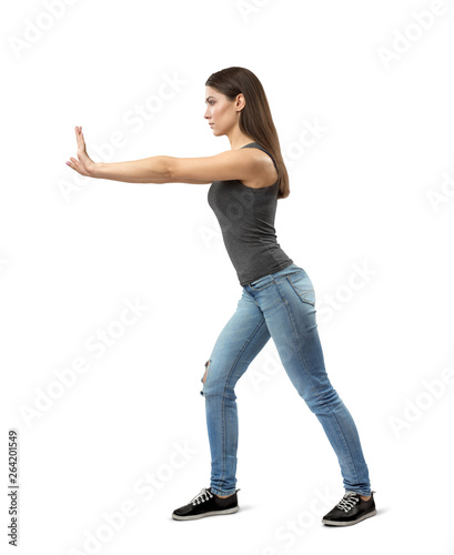 Side view of young fit woman in gray top and blue jeans, with long dark hazelnut hair, posing as if pushing something forward on white background.