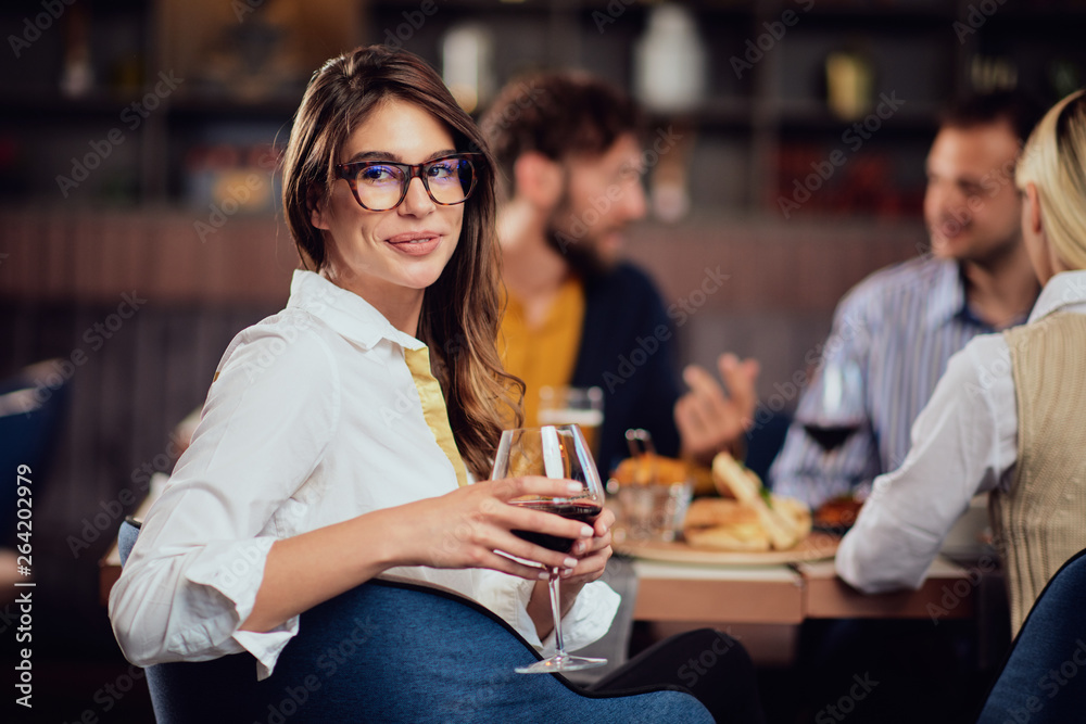 Portrait of gorgeous brunette looking at camera and holding glass with red wine while sitting at restaurat. In background friends eating diner. Portrait of gorgeous brunette looking at camera and holding glass with red wine while sitting at restaurat. In background friends eating diner.