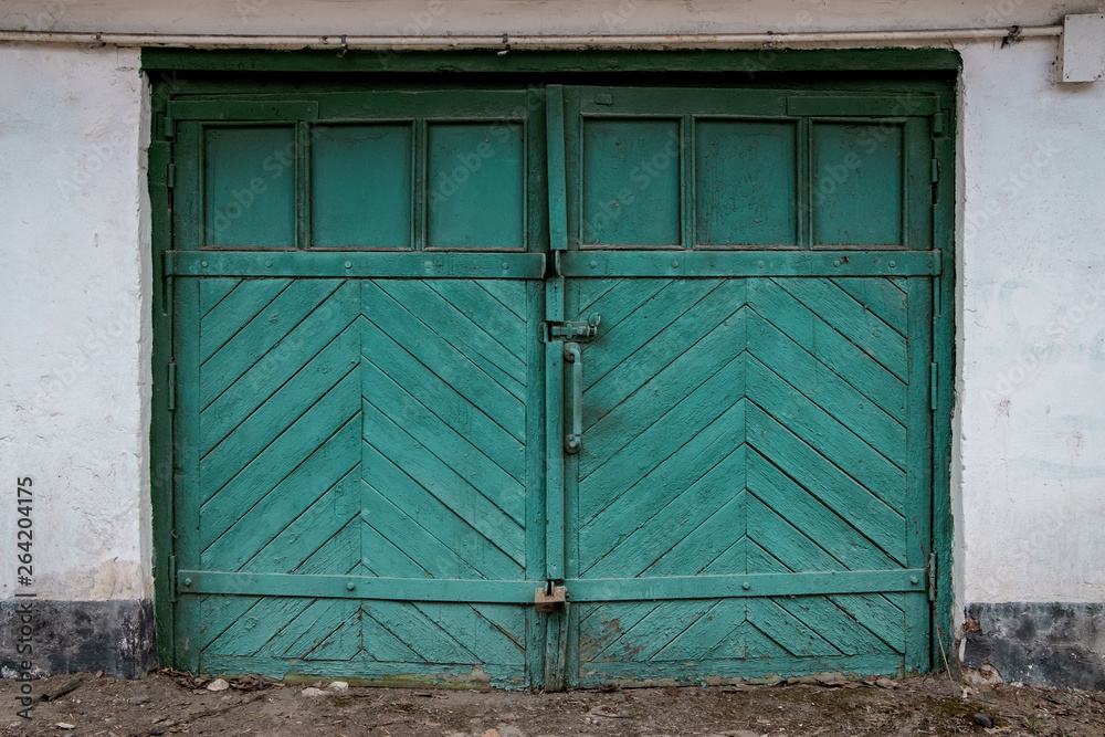 Old garage door in a white concrete wall. Gates made of wood painted with cracked green paint. Old garage door in a white concrete wall. Gates made of wood painted with cracked green paint.