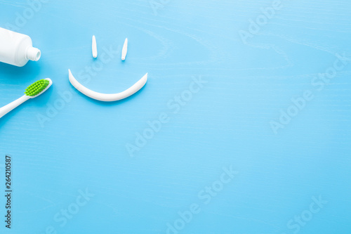 Toothbrush with green bristles, white tube of toothpaste on pastel blue background. Smiley face created from paste. Happy for healthy teeth concept. Empty place for text, quote, sayings or logo.  photo