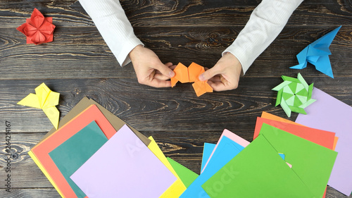 Hands making origami figure with orange paper. Top view. Wooden desk surface. photo