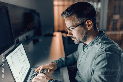 Bild auf Leinwand Serious Caucasian bearded architect with eyeglasses working on important project late at night in office