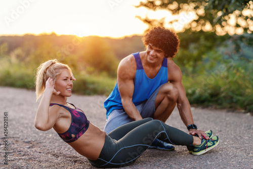 Gorgeous sportive blonde woman with ponytail doing crunches while her boyfriend holding her legs and helping her. Rural scene, summer sunny day.