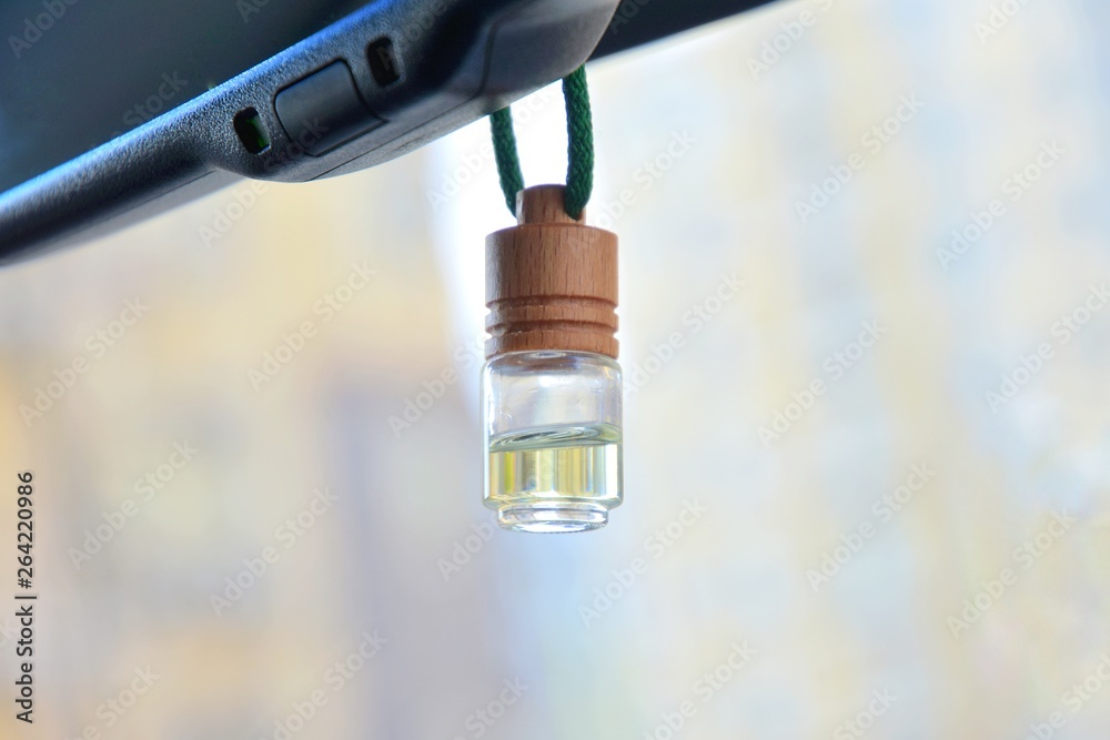 Car air perfume freshener bottle inside the car with part of car mirror on blurred city background. Little glass bottle with wooden lid and yellow aromatic liquid automobile freshener on a green rope.