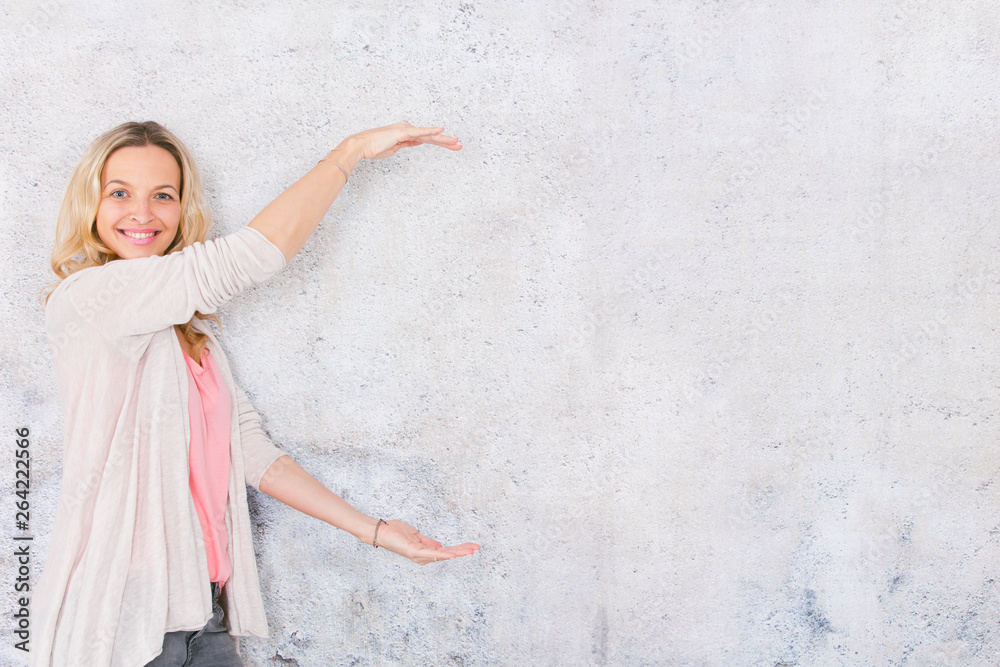 beautiful, blond woman is pointing at and posing in front of grey, concrete wall background beautiful, blond woman is pointing at and posing in front of grey, concrete wall background
