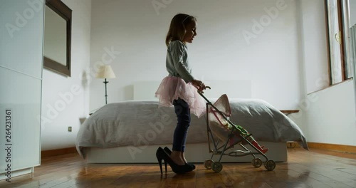 Authentic shot of little girl is playing with stroller toy and doll wearing her mother high heeled shoes in parents bedroom.