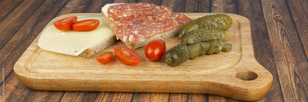 A slice of bread with salami and a slice of bread with cheese, while still small cocktail tomatoes and pickled cucumbers, served on a wooden board A slice of bread with salami and a slice of bread with cheese, while still small cocktail tomatoes and pickled cucumbers, served on a wooden board