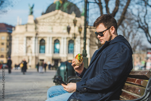 man eating burger surfing internet on phone sitting on bench
