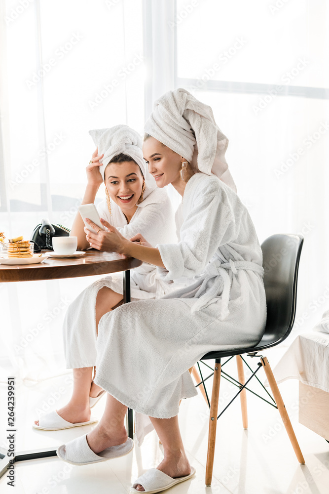 stylish smiling women in bathrobes and jewelry with towels on heads using smartphone during breakfast