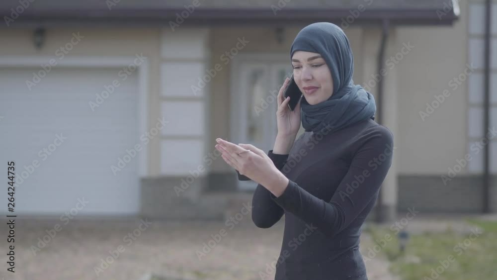 Young smiling eastern woman in balck clothes and beautiful headdress talking by cellphone holding keys in hand standing in front of big house. Asian woman in headscarf bought new home