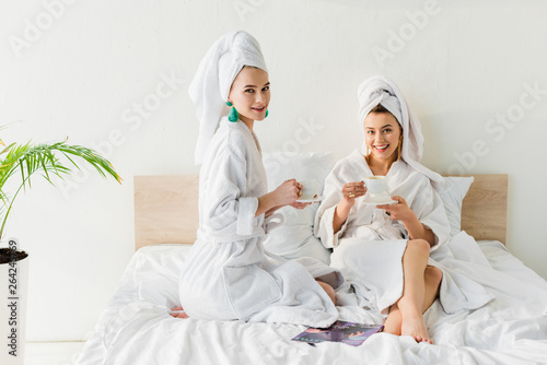 elegant women in bathrobes, earrings and with towels on heads looking at camera and holding coffee cups and saucers in bed