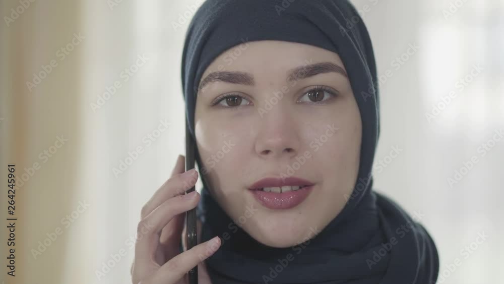 Portrait of a young beautiful eastern woman in the modern Muslim clothes and beautiful black headdress talking by cell phone close up. Young asian Muslim woman in head scarf looking in camera Portrait of a young beautiful eastern woman in the modern Muslim clothes and beautiful black headdress talking by cell phone close up. Young asian Muslim woman in head scarf looking in camera